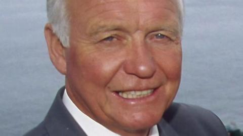 Craig Rich seen wearing a grey suit, white shirt and patterned tie. He is pictured with the sea in the background.