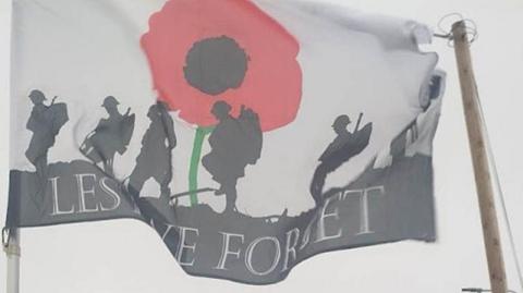 World War One soldiers in silhouette on a flag which has the slogan Lest We Forget. A large red poppy is in the background.