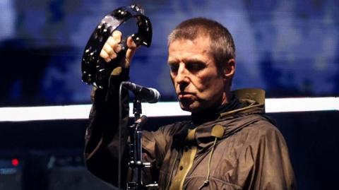 Liam Gallagher, wearing a raincoat, has a sombre expression as he holds up a tambourine during an Oasis reunion concert