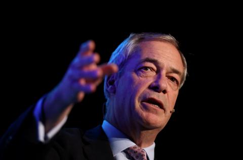 Nigel Farage speaks into a microphone to the side of his face as he point with his hand to something in front of him. He is wearing a suit and has grey hair.  