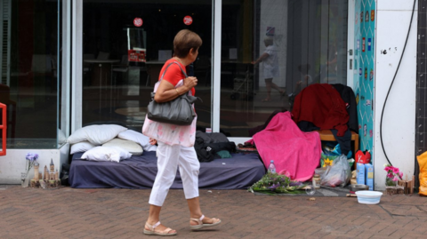 Woman walks past homeless person's mattress and belongings outside a shop.