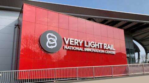 A large red panelled sign on the side of a grey building reads Very Light Rail National Innovation Centre in white lettering.
