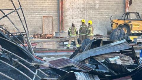 Fire fighters stand in a scrap metal yard with large sheets of metal lying around in piles. 