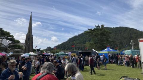 Stalls and crowds on the fair field at St John's. Manx flags are fling in the background, and the hill Slieau Whallian is to the right of the spire of the Royal chapel of St John's.