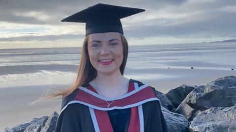 Francesca Murphy wearing graduation clothes and cap, standing, looking into camera with Swansea beach behind her. She is smiling.