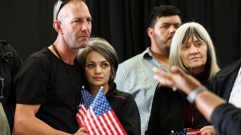 A man wearing sunglasses on his head with his arm around a woman. They are standing on the left and each holding a mini American flag. To their right, is an older woman. Behind her is a man. The background appears to be a black curtain.