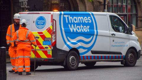 Two Thames Water workers wearing orange suits and white helmets stand at the back of a van with the company logo on the side. 