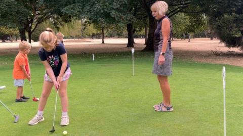Several people practice putting on a golf green surrounded by large trees and dry grass, with a child preparing to putt while others watch nearby.