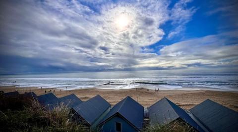 A row of beach huts look out on to a beach. A few people can be seen on the sand in the distance. The sky is a mix of clear blue, white clouds and more threatening grey clouds rolling in from the left