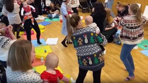 A group of women wearing Christmas jumpers standing in a circle holding babies