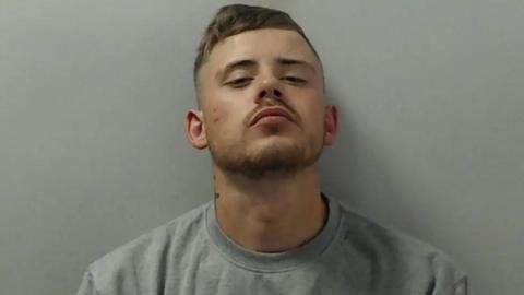 A mugshot of Liam O'Pray, wearing a grey sweatshirt. His head is tilted upwards as he looks down his nose at the camera.