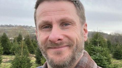 Chris Hurcombe, who has a brown and grey beard, is smiling while standing outside by trees. 