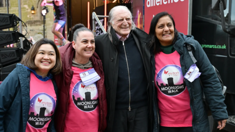 Three women posing for a picture with David Bradley. The women are dressed in pink shirts with Moonlight Walk logo on it. David, standing in the middle in a black zip up top and coat is smiling at the camera