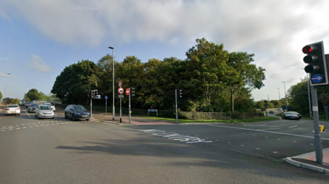 Street view image of the junction shows cars stopped at traffic lights. To the right of the cars are more traffic lights and a No Entry sign on the ground. 