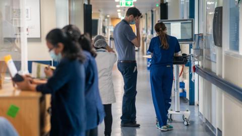 A group of five NHS workers engaged in tasks in a hospital corridor. Two women, dressed in blue uniforms, are examining documents while standing at a wooden desk. A woman wearing a white uniform has her back to the camera. A tall man dressed in a blue polo shirt and chinos stands by a computer station talking to a woman wearing a blue uniform.