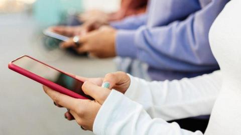 A close-up image of several young people using smartphones that appears to be indoor. The background is softly blurred, emphasising the hands and devices in the foreground.