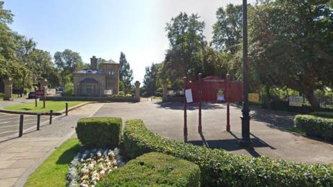 A gatehouse and a red sign saying Alexandra Park at the entrance, with a number of shrubs, cream coloured flowers and trees