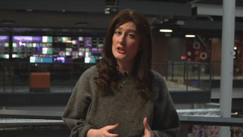 Gemma Dunstan at 91ȱ Cardiff Central square on a balcony in front of a tv control room. she has long dark hair and is wearing a loose grey top