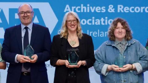 Three people stood in front of a screen at the awards ceremony. Catherine is stood at the centre and is holding an award, with two other people either side.