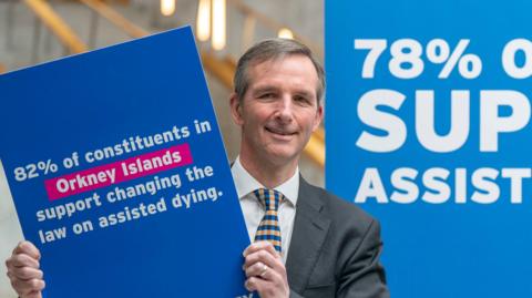 Liam McArthur - a man with brown hair and wearing a suit with a yellow and blue tie, holds up a placard saying people in the Orkney Islands support changing the law on assisted dying.