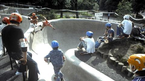 Young people in 1978 wearing helmets and other safety gear skateboarding at Kelvin Wheelies skatepark 