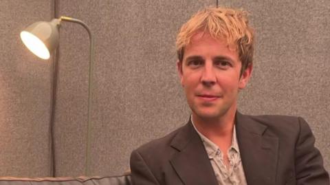 A head-and-shoulders image of Tom Odell sitting next to a lamp. He is smiling and looking into the camera.