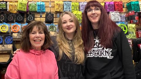 Three women. Dan's mum wears a pink hoodie and has short brown hair, his stepdaughter Scarlett has long blonde hair and is wearing all black. Dan's wife Ruth wears a black hoodie and has  long brown hair.