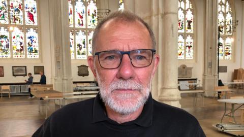 A picture of Matthew Porteous. He is standing inside St Edmundsbury Cathedral. Behind him pillars and stained glassed windows can be seen. Tables that will hold the model railway tracks are standing. He is facing the camera with an expressionless face. He has black glasses, dark thinning hair and a grey beard. He is wearing a black jumper with a black polo top underneath.