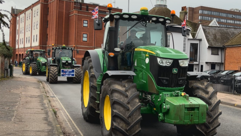 A green tractor with British flags on either side of the driver's cab moves through a town centre road. Several other tractors of similar colour and style follow it from behind. Buildings surround the road and there is a car park to the right hand side. 