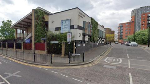 A road junction in east London. There are modern red apartment blocks at the end of the road and a modern school building at the junction. The number 20 indicating the speed limit is painted on a speed bump in the road. 