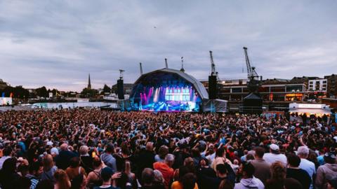 Lloyd's Amphitheatre in Bristol. The stage is a semi circle shape and it is lit up with pink and blue spotlights. There is a large crowd of people standing and watching the performance. The Bristol landscape can be seen in the distance behind the stage.