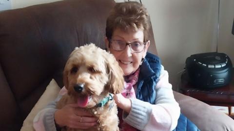 An older woman is sitting on a seat in a living room. She has short brown hair, wears glasses and a coat and is holding a dog.