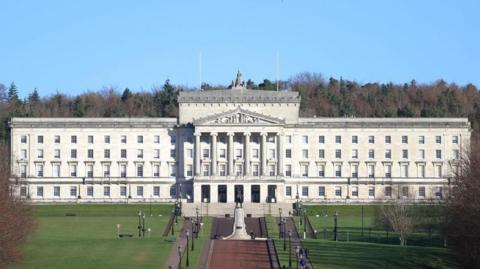 Stormont is a large stone building with stairs leading up to it. It is rectangular shape and has four rows of windows along the front, and a stone turrets at the front. On top of the building is a metal statue. Dotted around the grounds are black lampposts and benches for people to sit on. There is a statue feature in front of the steps to the front door.