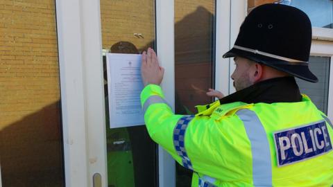 A police officer, wearing a high visibility coat and black helmet, is sticking a piece of paper, with the closure notice typed out, on a glass porch door.  