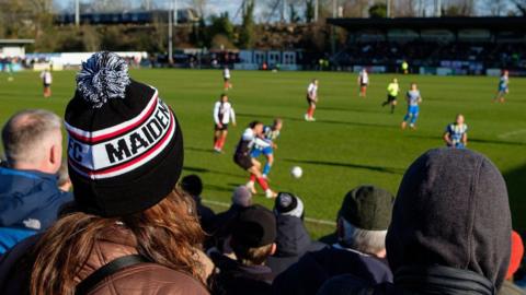 A crowd watch a football match. The players are out of focus in the background But you can see the backs of the crowds heads. They are mostly wearing coats and hats. The woman closest to the camera has long brown hair and is wearing a black and white bobble hat with the word Maidenhead on the side.