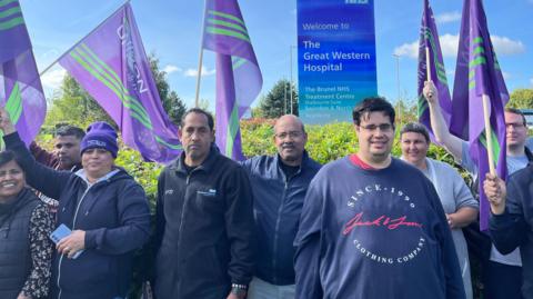 A group of eight people holding purple and green Unison union flags outside a hospital entrance sign that says Welcome to The Great Western Hospital.