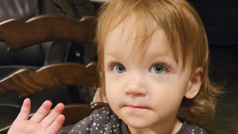 A two-year-old girl with shoulder-length brown hair and blue eyes is wearing a dark top with white stars on it. She is sitting on a chair and is holding up her right hand.