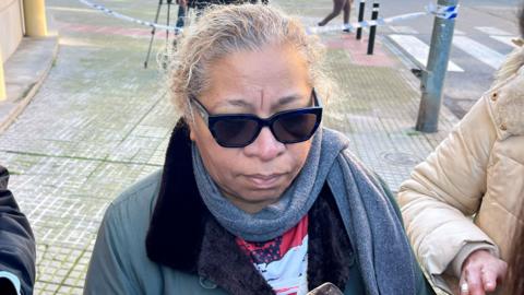 A woman in a grey scarf and coat talks to reporters