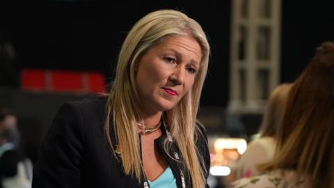 Laura Anne Jones speaking to an individual off camera, wearing a black jacket and blue top.