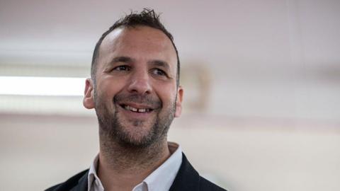 Zack Polanski looking to the side wearing a white shirt and black blazer with white backdrop