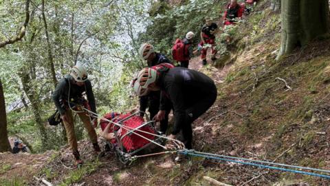 An injured man is lying on a stretcher, which is being pulled up a hill using ropes and a pulley. Four mountain rescuers in white helmets surround the man to assist him, while other rescuers stand on the path below. They are all in a wooded hilly area.