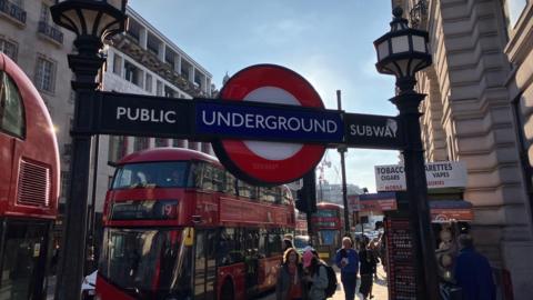 In the foreground is a blue and red Tube roundel between 'Public subway' sign. In the background is a red double decker 19 bus and people walking along the pavement