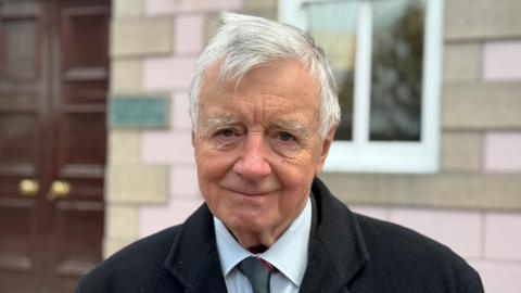 Deputy Philip Bailhache smiling at the camera. He is wearing a black coat, a white shirt and silver tie. He is stood in front of a building with light brown and pink bricks.
