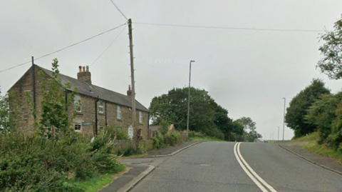 Lobley Hill Road, at the junction of Schoolhouse Lane, on the A692. There is a cottage to the left of the double carriageway road.