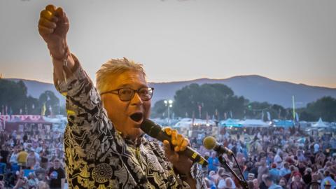 A man with grey hair is facing the camera and is shouting into a microphone on a stage, with his one arm raised in the air. Behind him is a huge crowd of people, and beyond them are lit-up tents, trees and rolling hills