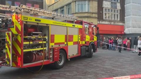 A yellow and red fire engine parked outside a shopping centre and behind a cordon of red and white tape which says 'do not cross'. Bystanders are behind the cordon watching on.