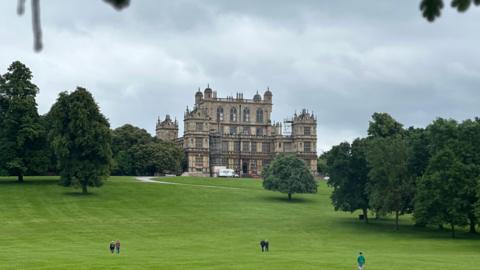 A grand stately home on the top of a sweeping green hill surrounded by trees