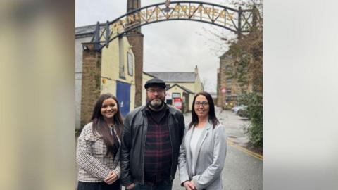 Chris France (middle) with flat cap and beard and leather jacket and Rebecca Canfield (right) with grey cardigan and glasses and dark brown hair. There is a woman to the left of the pair wearing a chequered jacket. 