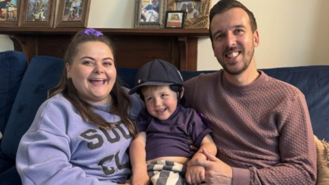 A toddler sat in between his mum and dad on a sofa, all smiling 