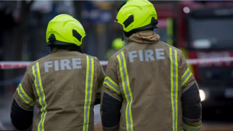 The back view of two firefighters - both wear brown jackets with green and grey lines on the back and the word "fire" emblazoned on them. Both wear green helmets.